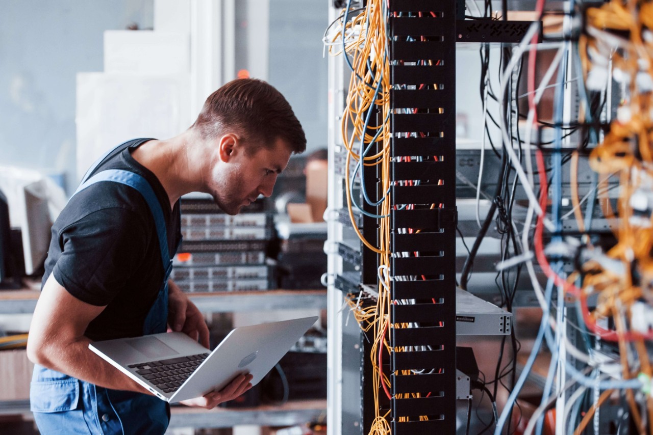 Young man in uniform and with laptop works with internet equipment and wires in server room Young man in uniform and with laptop works with internet equipment and wires in server room